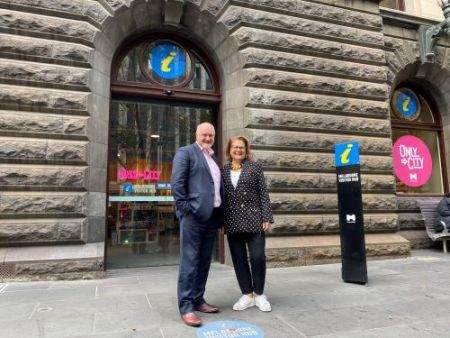 Pictured outside the Melbourne Visitors hub, a man in a suit hands in pockets and a woman in a black white spotted jacket. The building is grey historical stone. A blue logo with a yellow i is on two signs.