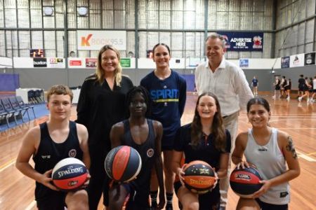 Three people standing - a woman local MP, a woman Bendigo Spirit basketball player, a male TAC representative. Four people kneeling in front of them holding basketballs - two male basketball players and two female. The photo is taken in a basketball stadium. 