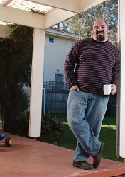 A middle aged man leaning against a verandah post while holding a mug.