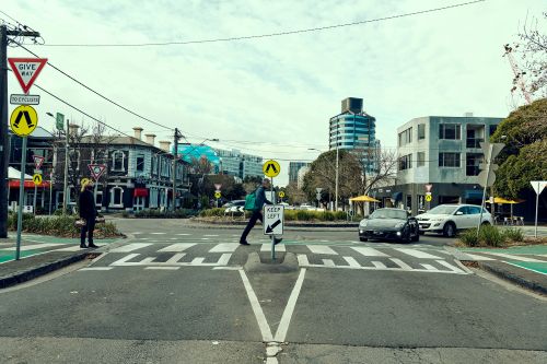 A raised pedestrian crossing in a city. One person is walking across the crossing another waiting to cross. Cars are parked waiting for the people to cross. 