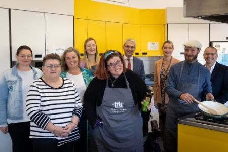 9 people standing looking to camera in a kitchen.