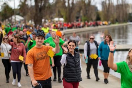 A large group of people walking beside a lake smiling and waving to the camera. Two people in the foreground, one holding a guide dog lead. 