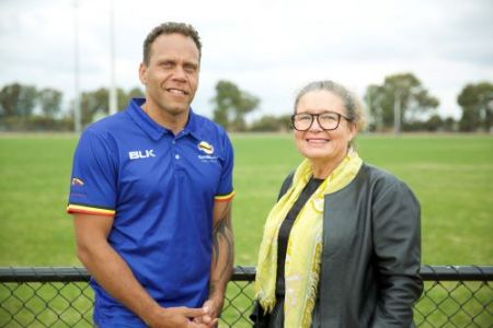 Two people looking to camera standing in front of a fence near a football oval.