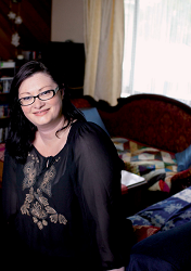 A dark haired woman with glasses sitting on the edge of a couch.