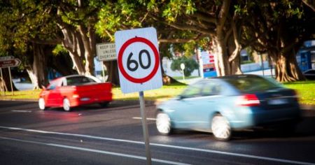 A road sign with 60 in a red circle. Two blurred cars on a road behind it. 