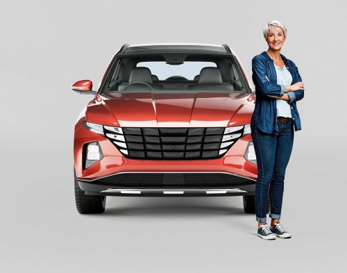 Woman with grey short hair, blue jacket, jeans and runners standing smiling with arms crossed in front of a red modern SUV. 