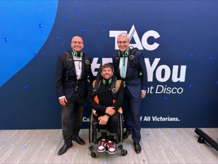 Three people in front of a TAC back drop. Two standing behind person in a wheelchair. All wearing headphones around their necks and haptic vests 