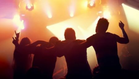 Four people arms around each other facing away from the camera towards the lights of a stage performance