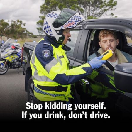 A policeman wearing a motorbike helmet holds a breath testing device while testing a male driver in a black ute. A police motorbike is in the background.