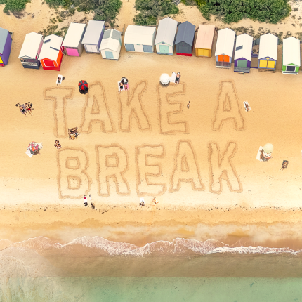 A image looking from above to the beach with sand and the ocean meeting. Several colored bathing boxes are in a row at the top of the image, a few people under umbrellas on the sand and the words Take a Break written in the sand. 