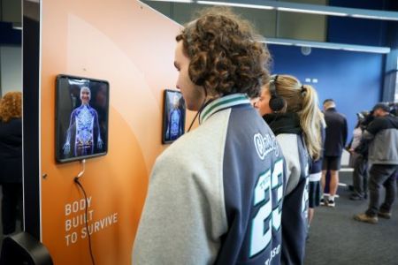 A school student wearing headphones looking at a small screen on a wall showing his face on a interactive drawn body. 