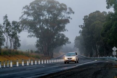 White car driving on a country road with gum trees and fog in the background. Flexible barriers are on the side of the road.
