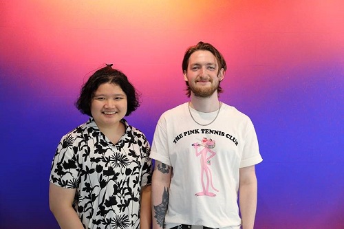 Woman in white shirt with black pattern, dark hair. Man with beard in white t shirt. Background purple and pink colours.