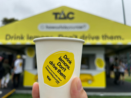 A white paper cup being held up in front of a yellow tent. The cup has drink lemons, don't drive them written ion it in black text inside the picture of a lemon. The tent in the back is blurred and has the TAC logo and how safe is your car website address written on it. 