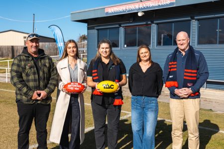 Standing in front of the Southside Junior Football club are four people - man wearing cap with green checked jacket and black pants, woman in long white coat, and blue jeans holding an AFL football with Be the Difference written on it, woman in black wearing a Southside footy scarf holding a yellow football with TAC written on it, a woman in a black shirt and jeans, a man wearing a blue top, beige pants wearing a Southside footy scarf. 