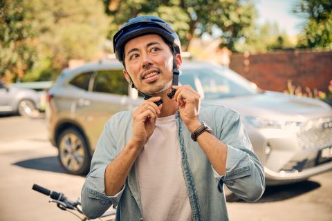 Man puts on bicycle helmet, standing beside a bike with a car blurred in the background. 