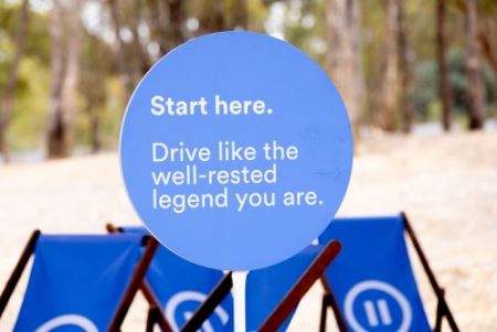 A round blue lollypop sign with the words in white. Start here. Drive like the well-rested legend you are. In the background - trees and dirt ground