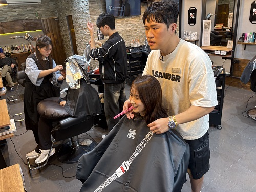 A haircutting salon with a haircutting standing behind a woman wearing a haircutting cape with a seatbelt across it. 