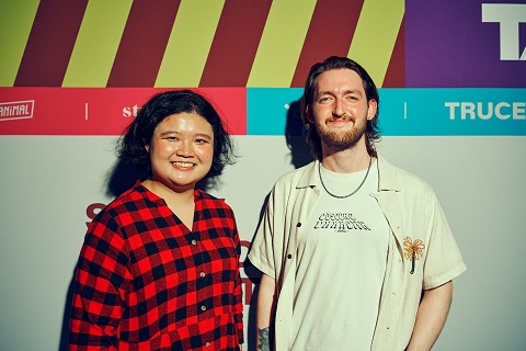 A woman with dark hair, wearing a red and black checked stands with a man, bearded, wearing a cream t short and shirt. Both are in front of a coloured backdrop with Split Second Competition branding. 