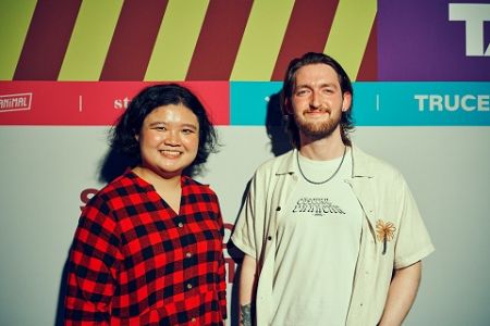 A woman with dark hair, wearing a red and black checked stands with a man, bearded, wearing a cream t short and shirt. Both are in front of a coloured backdrop with Split Second Competition branding. 