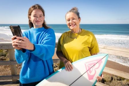Woman in blue jumper holding a phone next to a woman in a yellow dress holding a surfboard with the ocean in the background. 