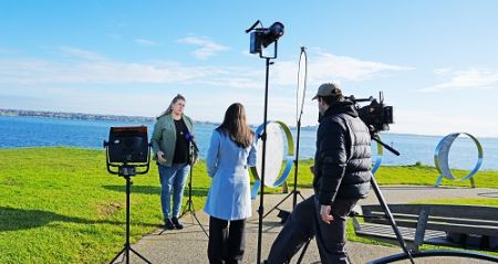 An interview set up with the ocean and green grass in the background. A man in a black puffer stands beside a camera, a woman in a long blue jacket back to camera interviews a woman wearing a green jacket, black top and jeans standing before a microphone. Lights are set up facing the interview.
